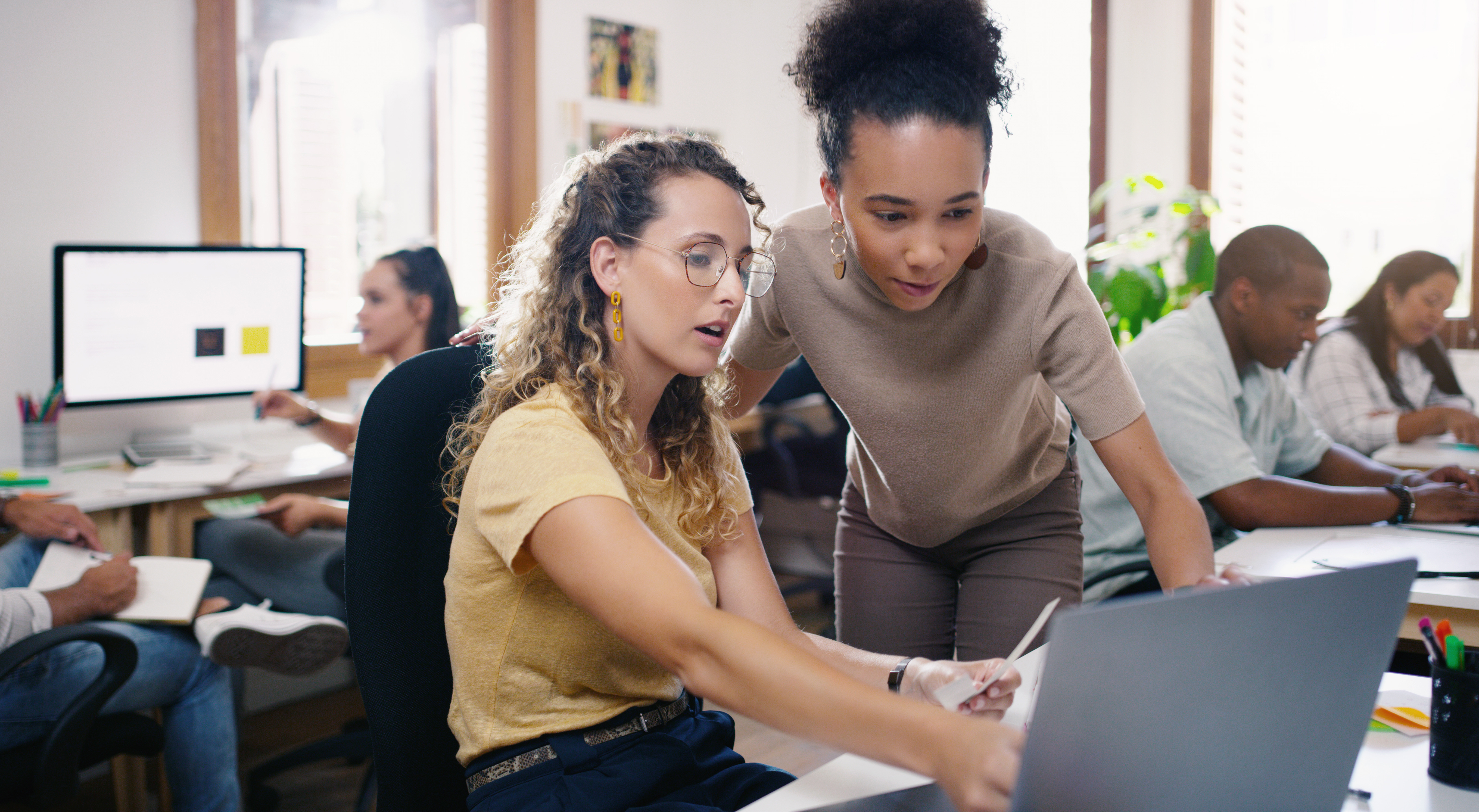 Im Hintergrund arbeiten mehrere Menschen an ihrem Schreibtisch. Vorne sieht man zwei junge Frauen, die gemeinsam auf einen Laptopbildschirm schauen und sich angeregt unterhalten. 