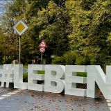 Auf einer Landstraße kommt von vorne links ein Polizeifahrzeug mit eingeschaltetem Blaulicht entgegen. Auf der rechten Seite stehen in einer Straßeneinmündung die lebensgroßen weißen Buchstabenblöcke #LEBEN.