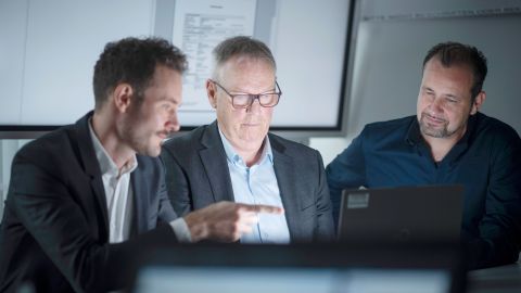 Dennis Sopp (LZPD NRW), Jürgen Marten (LKA NRW) und Michael Richter (PP Aachen) sitzen in einem Büro gemeinsam vor einem Laptop und schauen auf den Bildschirm. Dennis Sopp zeigt mit dem Finger auf den Bildschirm. Im Hintergrund ist eine weiße Wand sowie ein großer Bildschirm zu sehen.